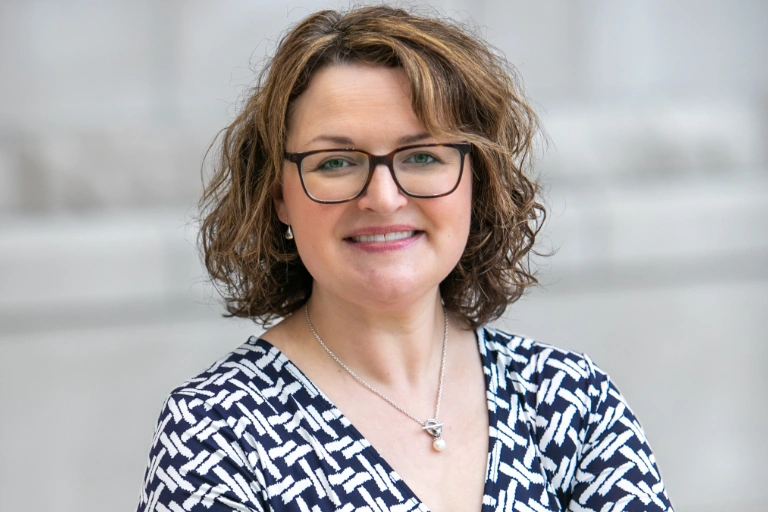 Image of a woman with short curly hair and glasses, wearing a black and white dress, smiling at the camera.