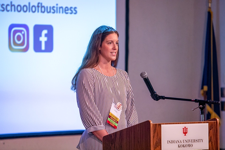 A woman with long brown hair, wearing a nametag with yellow, crimson, green, and red labels and a blue and white pinstriped blouse speaks at a podium, in front of a presentation shown on a large screen.