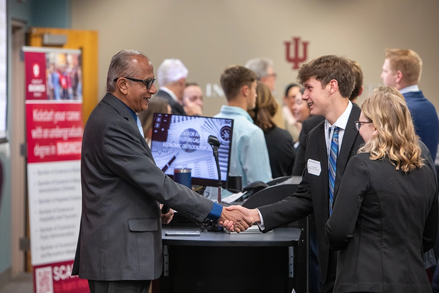 On the left of the image, an older Indian man wearing glasses and a gray suit shakes the hand of a younger man in a blue suit, white shirt and blue patterned tie.