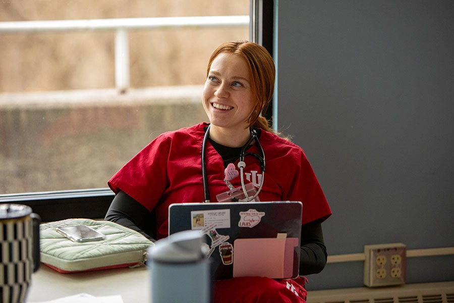 A female student in red scrubs with the IU trident over a long-sleeved black shirt smiles as she works on a laptop.