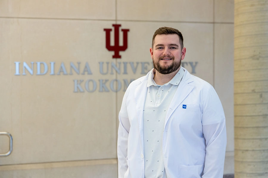 A man in a medical white coat and patterned polo shirt stands and smiles for a photograph in front of a wall featuring the words Indiana University Kokomo underneath the Indiana University trident.