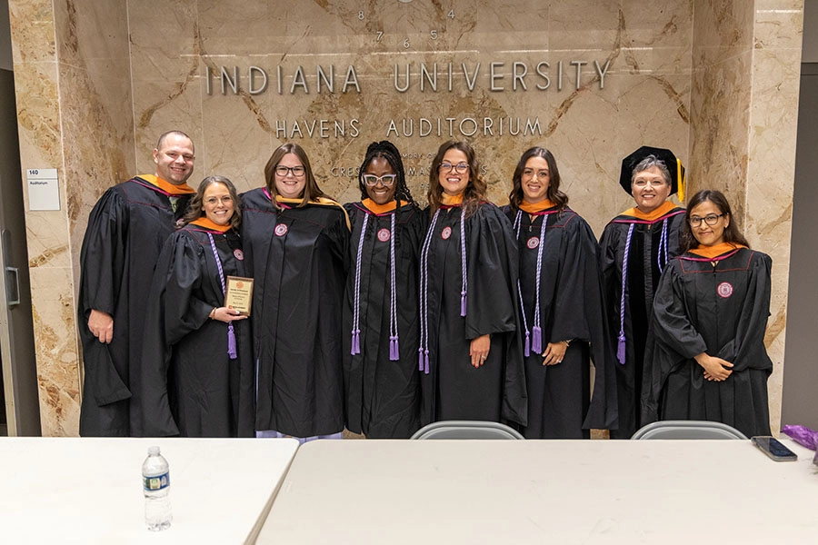 One man and seven women wearing black commencement robes and apricot hoods pose for a photograph in front of a wall bearing information about Indiana University Kokomo's Havens Auditorium. Five women are also wearing purple cords, one is wearing a doctoral tam.