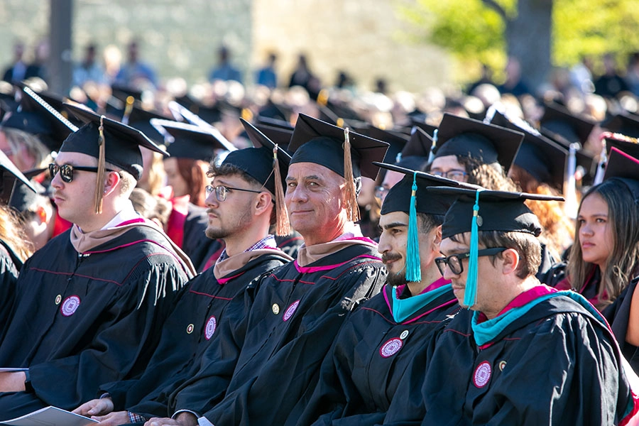 An older man wearing a black commencement cap with gold tassel, black commencement gown, and drab masters hood looks sideways at the camera and smiles, while surrounded by men and women in black commencement gowns and caps, along with multiple colors of hoods and tassels.