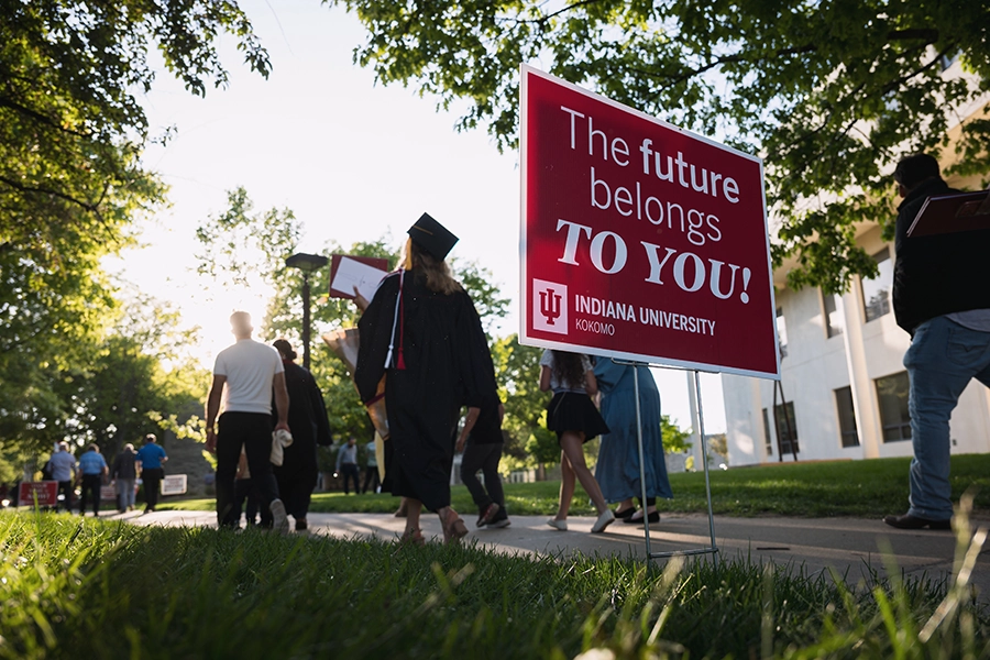 People, some wearing graduation caps and gowns, walk past a yard sign with the words 'The future belongs TO YOU'. The sign also features the logo for Indiana University Kokomo and the Indiana University crimson trident in a white box.