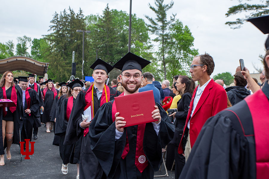 A man in a commencement cap and gown holds a red diploma cover.