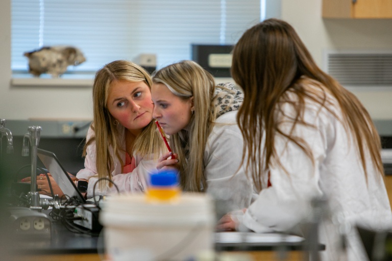 Three women in lap coats looking at a laptop.