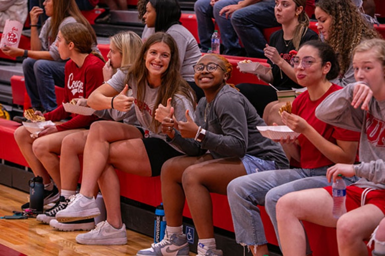 A group of students sit on bleachers. Two women smile and give thumbs up to the camera.