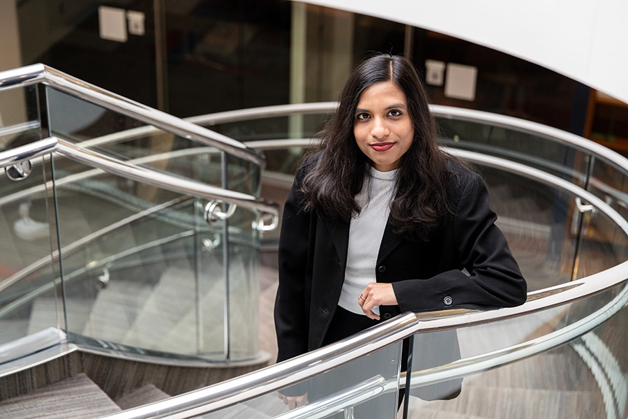 An Indian woman with black hair, wearing jeans, a gray turtleneck and a black blazer, stands on a circular staircase and looks up at the camera.