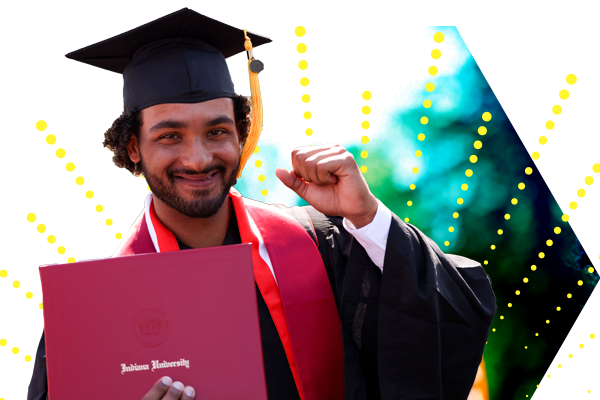 a male graduate in cap and gown holds his crimson IU diploma in one hand with his other in the air