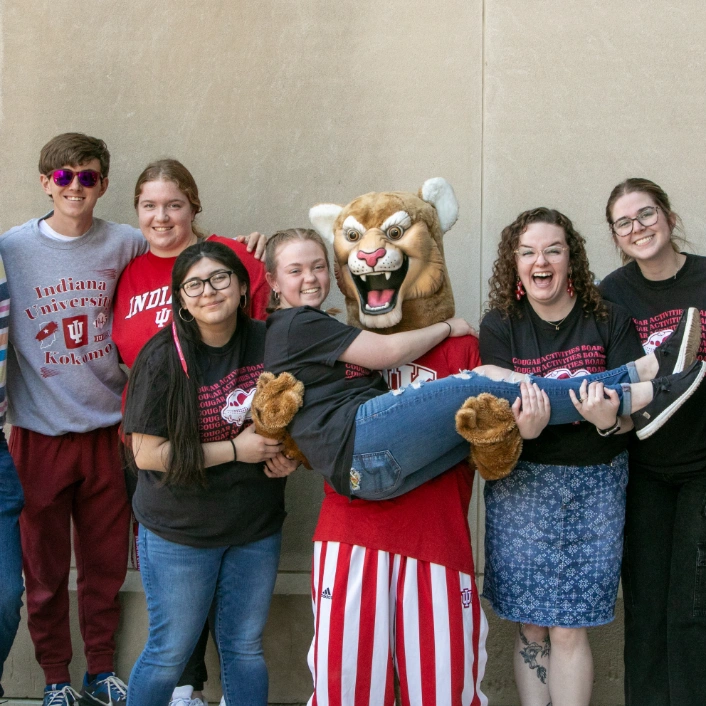 A group of men and women smile for the camera. One woman is held by a person in a Cougar mascot suit. 