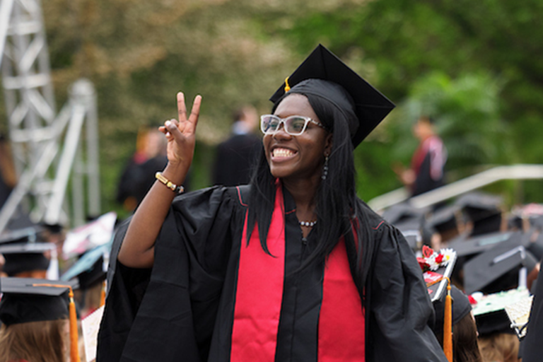 Female undergraduate student in commencement robes throwing up a peace sign while walking up to accept her diploma