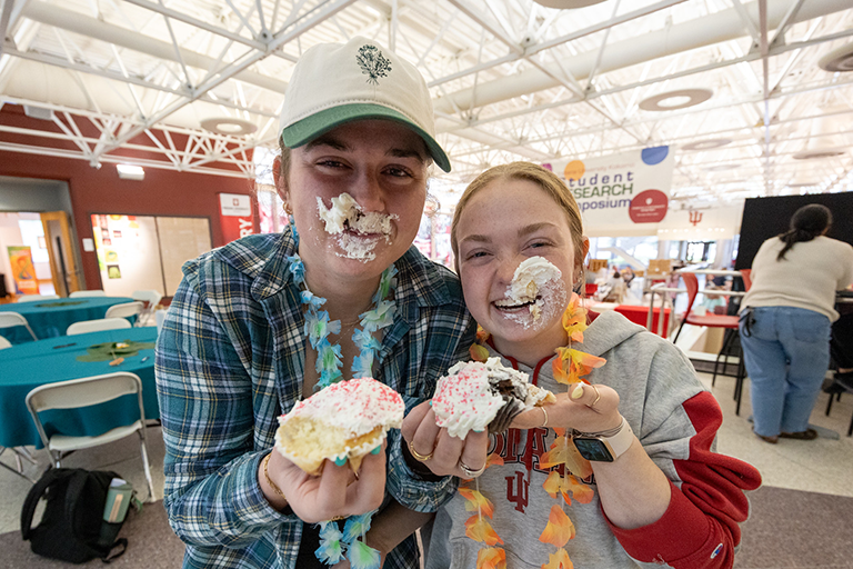 Two students eating their cupcakes, the icing is all over their faces and they are laughing toward the camera showing their pastries.