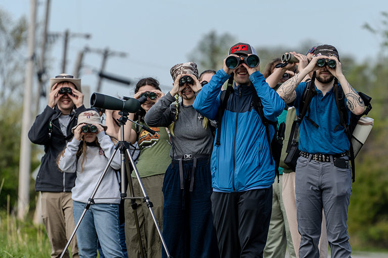 a group of 10-15 students all looking at the camera with binoculars at the Indiana Dunes State Park.