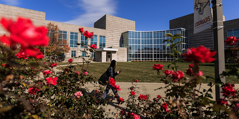IU Kokomo campus, foreground is full of pink and red flowers, middle ground has students walking along a path in front of a main campus building on a sunny day.