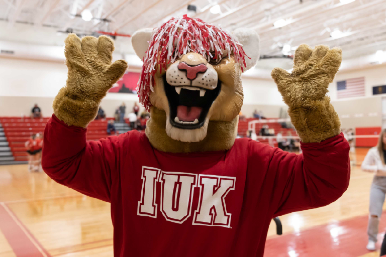 The IU Kokomo cougar mascot poses on a basketball court with its arms raised and a pom pom on its head.