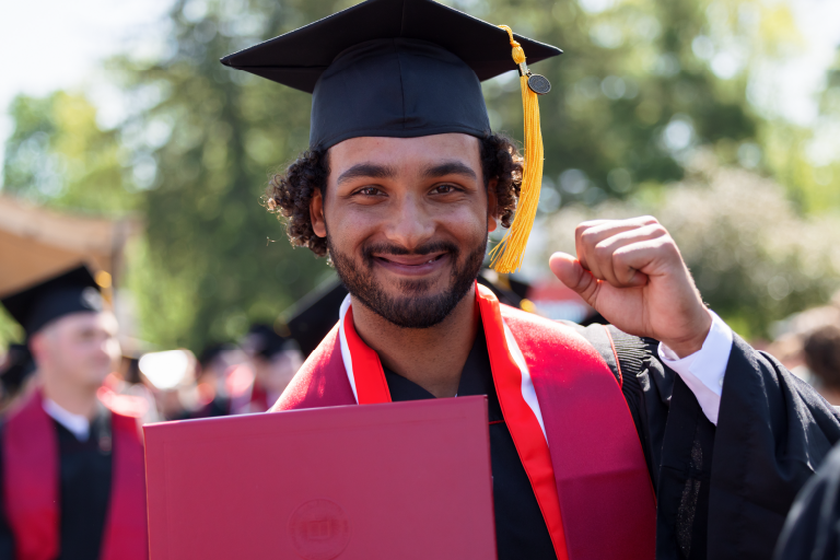 A male student with curly hair wears a cap and gown, has his fist raised in celebration, and holds his diploma.