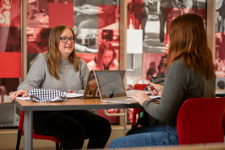 Two female students sit across from one another at a table with their laptops open, engaged in conversation.
