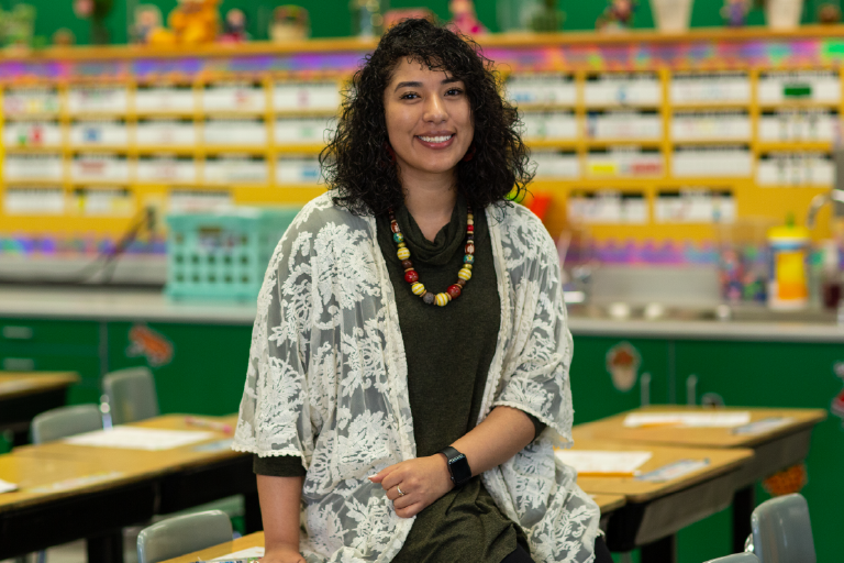 A woman smiles at the camera while leaning against a desk in a classroom with rows of desks.