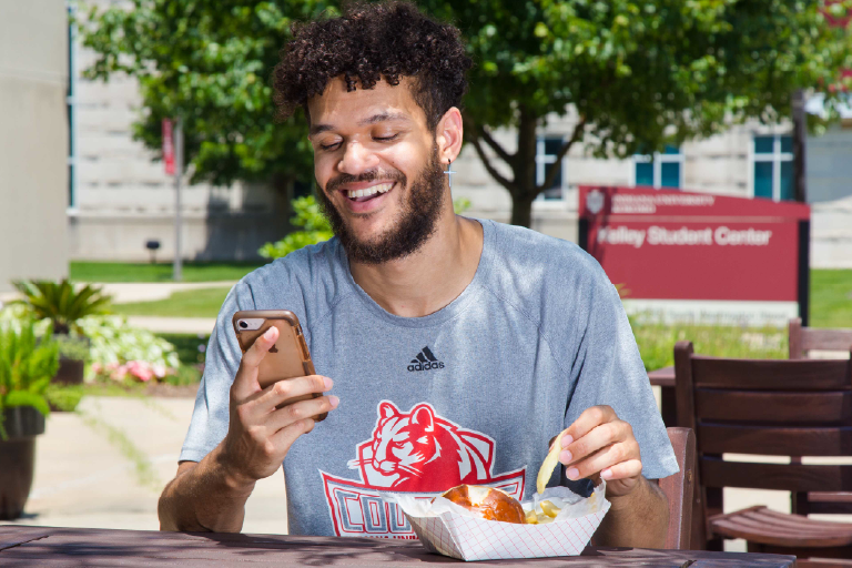 A male student wearing an IU Kokomo t-shirt sits outside at a table, holding his phone while smiling.