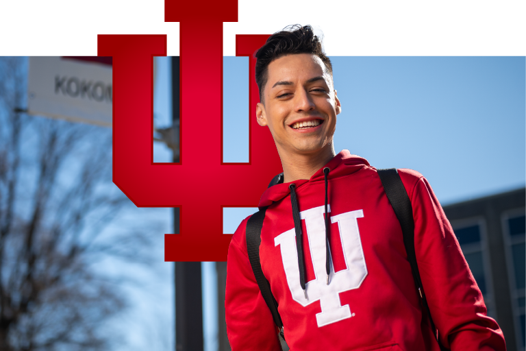 A male student wearing a red IU sweatshirt and a backpack poses in front of a building at IU Kokomo. The IU trident is superimposed to his left.
